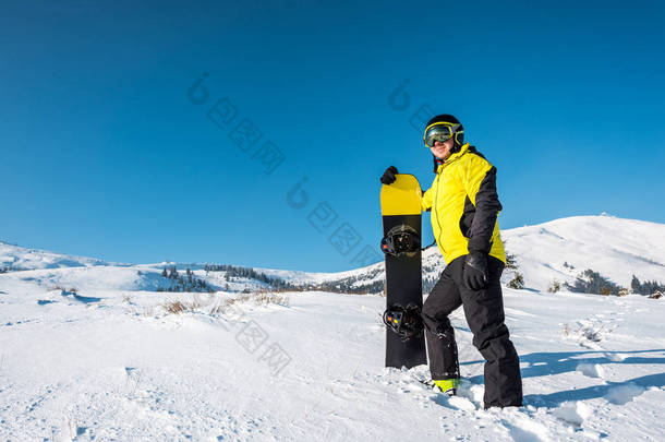 戴着头盔的快乐运动员拿着雪板站在山上的白雪上