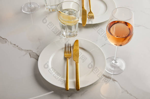 玻璃杯旁边的白盘上有金制刀叉，玻璃杯上有玫瑰酒，大理石桌上有柠檬水