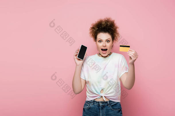 兴奋的女人拿着用粉红隔离的<strong>信用卡</strong>和智能手机