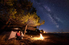 在海边过夜露营。徒步旅行的妇女准备食物在一个气体燃烧器, 休息在旅游帐篷入口在森林附近对明亮的蓝色星光天空与银河