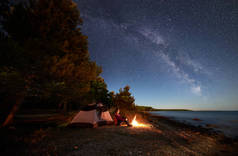 在海边过夜露营。女徒步旅行者坐在帐篷前的篝火旁, 在明亮的星空和银河下, 享受着美丽的蓝色水景色。旅游与积极的生活方式理念