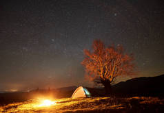 梦幻般的夜露营网站视图。明亮的篝火在美丽的星空下在旅游照明帐篷附近燃烧。大树和远处山脉的背景。旅游业和旅游概念.