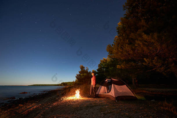 在海边过夜露营。徒步旅行的妇女休息在篝火旁的旅游帐篷和森林在星空<strong>下</strong>, 享受清澈的<strong>蓝色</strong>水的美丽景色。旅游与积极的生活方式理念.