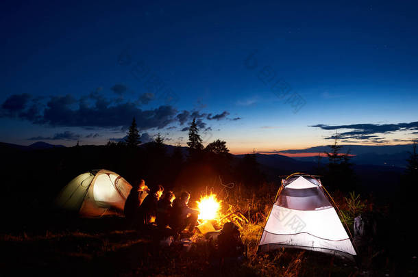 家庭徒步旅行的母亲, 父亲, 两个孩子在山上露营休息, 坐在篝火旁的日志和两个照明帐篷, 享受夜空, 日落, 星星惊人的景色