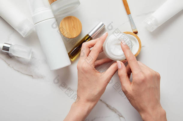 在白色表面用眼刷握住奶油罐的妇女的裁剪视图，即靠近管子、化妆品分配器和玻璃瓶