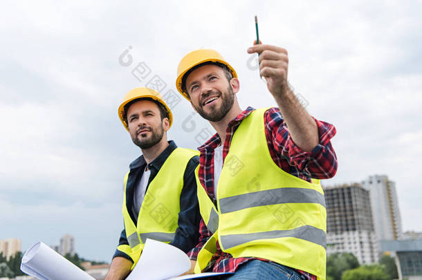 两名建筑师在<strong>安全背心</strong>和 hardhats 工作的蓝图和指向建筑