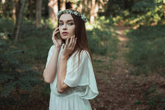 美丽的温柔的女孩摆在白色礼服和花卉花圈在森林里