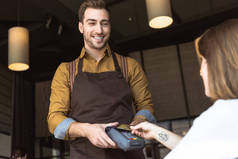 微笑的年轻侍者持有付款终端, 而客户做非接触购买与信用卡在咖啡厅
