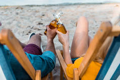 在沙滩上坐在太阳椅上的情侣的裁剪图像和啤酒瓶的玻璃杯