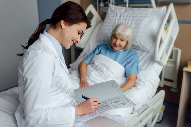微笑的女医生坐在床上写诊断, 而老年妇女躺在床上<strong>住院</strong>
