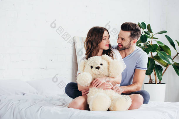 幸福的夫妇拥抱泰迪熊, 而坐在床上与白色的床上用品