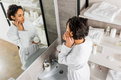 非洲裔美国妇女应用化妆品防皱霜面部和镜子在浴室里寻找