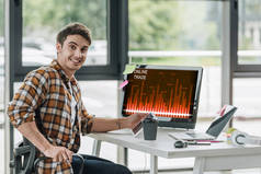 微笑的程序员看着相机，而坐在电脑显示器附近与屏幕上的在线交易
