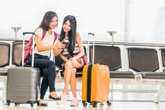 两个年轻的亚洲女孩使用智能手机检查航班或网络登记, 坐在机场等待座位在一起。空中旅行的生活方式, 令人兴奋的暑假旅行或手机小工具应用概念