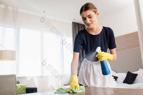 女服务员穿着围裙和橡胶手套，拿着喷雾器和抹布在宾馆房间清洁木制表面