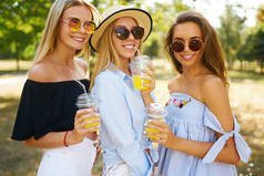 三个年轻貌美的姑娘在炎炎夏日玩得开心,喝着新鲜的果汁.朋友们在公园的背景下享受橙汁和摆姿势。夏季概念.