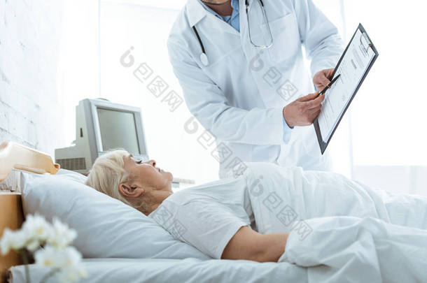 cropped 视图医生与剪贴板和生病的老年妇女躺在床上