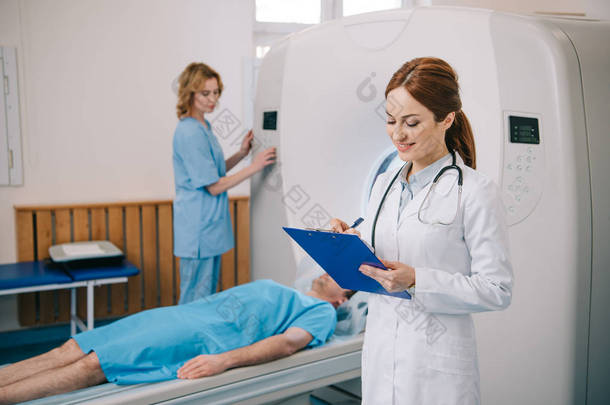 微笑<strong>放射</strong>科医生在剪贴板上写作的选择性焦<strong>点</strong>，而助理操作ct扫描仪附近的病人躺在ct扫描仪床上