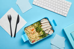 午餐盒的顶视图与美味的米饭与鸡肉和花椰菜在工作场所与计算机键盘的蓝色背景