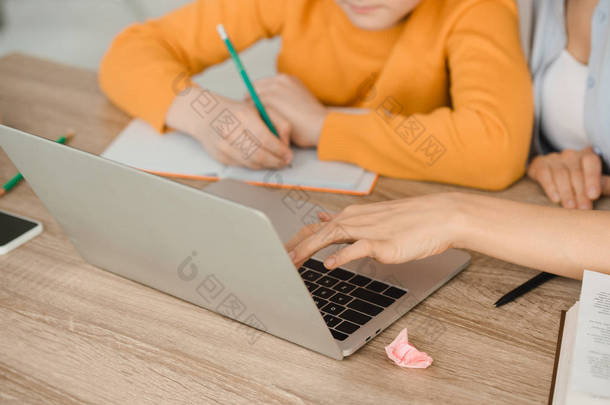 在家做功<strong>课时</strong>，使用笔记本电脑和男孩在抄本本上写作的妇女的裁剪视图