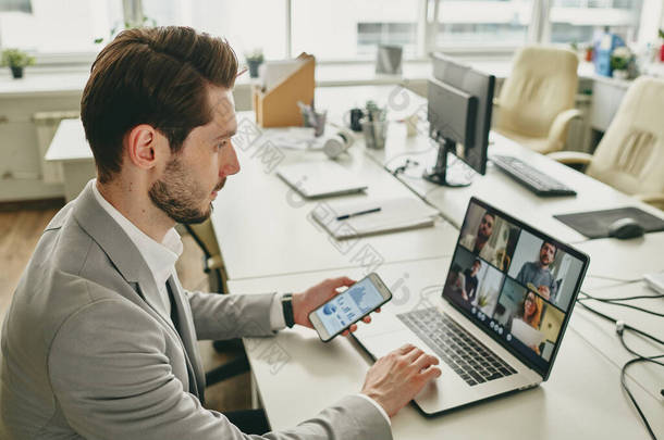 年轻的大胡子商人一个人在办公室里工作，通过笔记本电脑上的视频聊天将同事联系在一起