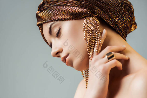 年轻的裸体女人，紧闭的眼睛，闪亮的妆容，金戒指和耳环，戴着头巾，摸着灰色的脖子