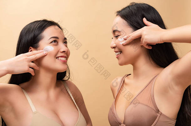两个开朗的多元文化的女孩应用在脸上的化妆品奶油和微笑隔离米色