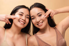 两个快乐的多元文化的妇女应用化妆品霜在脸上, 看着相机孤立的米色