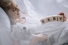 微笑的孕妇在白色睡衣与泰迪熊和木块与婴儿刻字在沙发上休息的侧面视图
