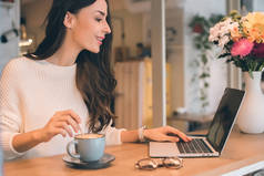 美丽的女性自由职业者的侧面视图工作在笔记本电脑在桌子上咖啡杯在咖啡馆