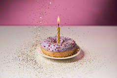 在甜甜圈中间燃烧蜡烛, 在粉红色背景的白色桌子上落下金色的火花