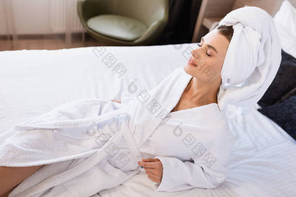快乐的女人，紧闭双眼，裹着毛巾，身穿白色浴衣，躺在酒店房间的床上