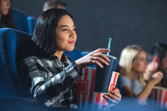 微笑亚洲妇女的侧面看与爆米花和苏打饮料观看电影在戏院