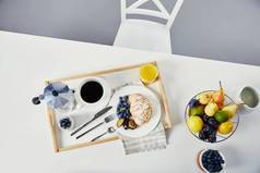 在白色桌面上的木托盘早餐的新鲜蓝莓和李子片, 玻璃汁, 杯咖啡和酸奶的顶部视图