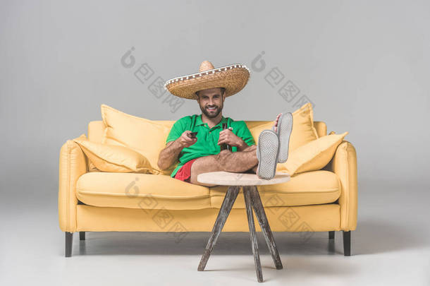 愉快的人在墨西哥草帽用瓶啤酒看<strong>电视</strong>在黄色沙发上<strong>灰色</strong>