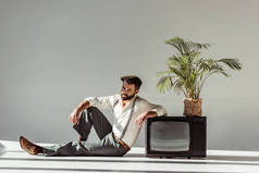 英俊的胡须男子在帽子坐在地板上附近的老式电视与植物在锅里, 看着相机