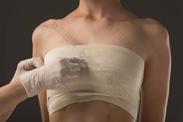 乳胶手套中整形外科医生的部分视图和在灰色上隔离的乳房绷带患者