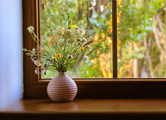 Bouquet of different wildflowers in a pink vase on a wooden window sill against a window at summer s