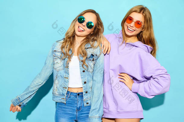 两个年轻漂亮的金发碧眼的女孩穿着时髦的<strong>夏装</strong>。性感的无忧无虑的女人摆在太阳镜的蓝色墙壁附近。积极的模型疯狂