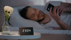 Clock and rose on bedside table near blurred man using smartphone on bed at night