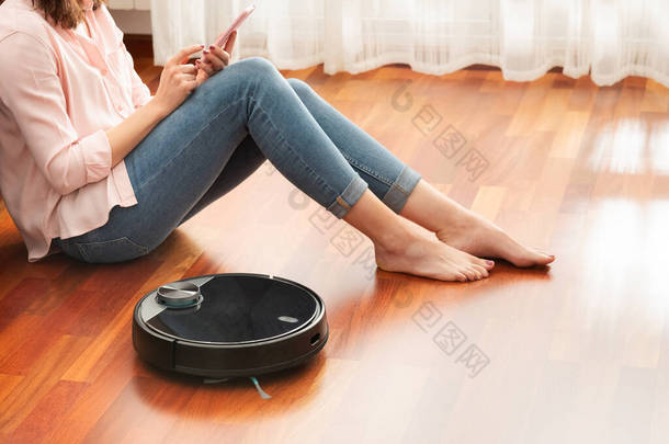 无脸中段的年轻女性使用自动真空吸尘器清洁地板，用智能手机控制智能机器家务机器人