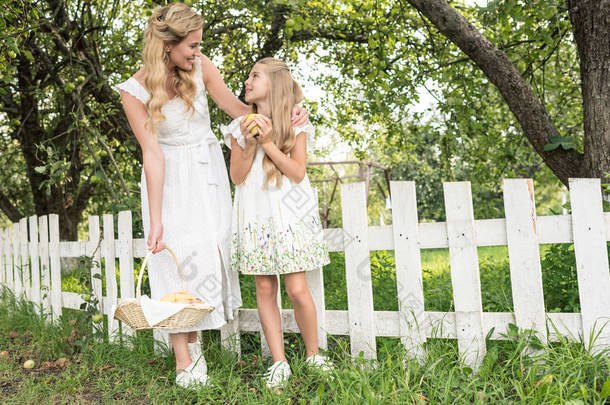 金色的母亲和女儿与水果在柳<strong>条</strong>篮子在花园附近的白色围<strong>栏</strong>摆