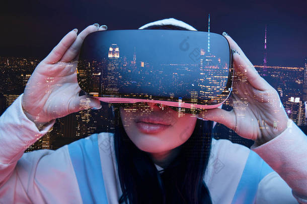 黑发女孩触摸虚拟现实耳机和现代城市与摩天大楼在夜间的双重曝光
