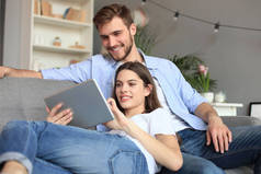 一对年轻夫妇坐在客厅的沙发上，坐在平板电脑上在线观看媒体内容.