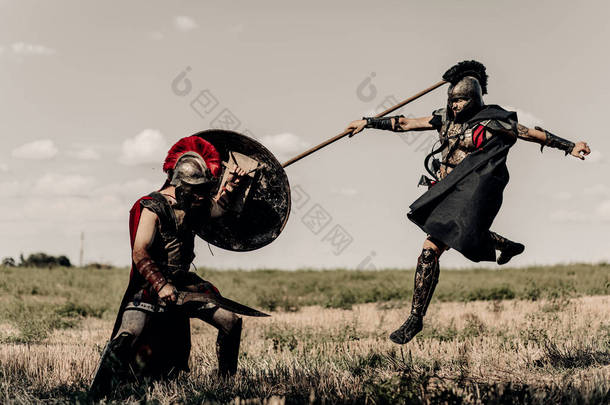 两个身穿战斗服的古希腊战士或罗马战士，在天空的衬托下，在草地上，用矛和剑战斗.