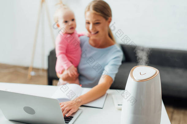 用笔记本电脑和抱着女婴的妇女身边<strong>加湿</strong>器的选择性聚焦