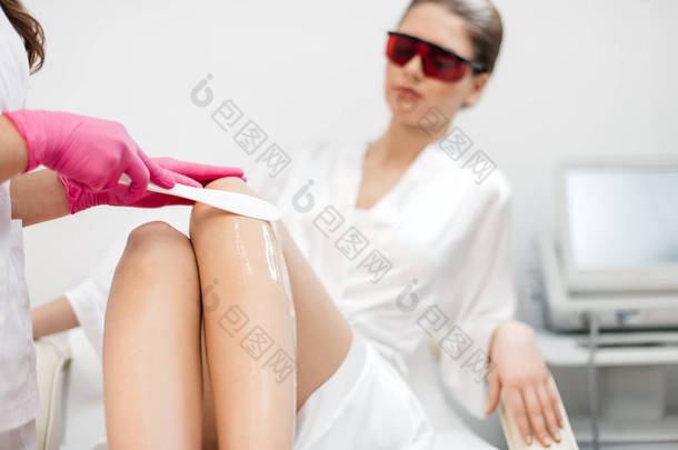 脱毛在美容院。美容师正在应用奶油为脱毛使用的蜡棒在迷人的女人的腿上, 红色安全护目镜在模糊的背景下.