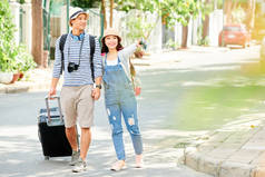 漂亮的年轻亚洲妇女握着男朋友的手与大手提箱，并指着酒店，他们正在寻找