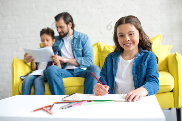 可爱的<strong>孩子绘图</strong>与彩色铅笔和微笑的相机, 而父亲和弟弟坐在沙发后面
