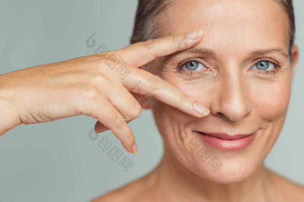 微笑的资深妇女的肖像与完美的皮肤显示胜利标志近眼睛在灰色背景。特写脸的成熟妇女显示成功后, 抗衰老的皱纹治疗结果。美容成熟肌肤护理理念.